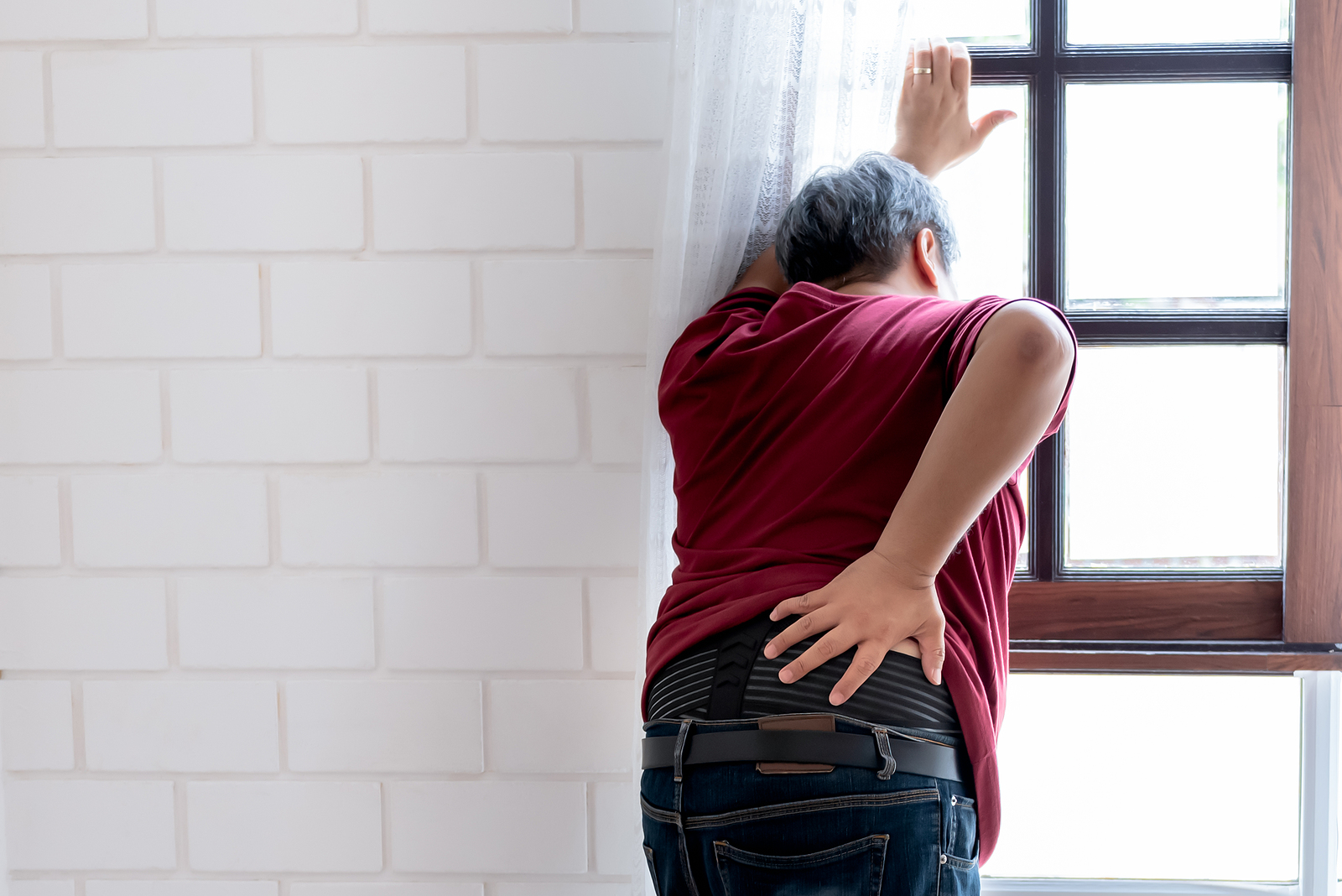 A man stands hunched over with a hand on their back while looking out a window. This could symbolize back pain that pelvic pain therapy in Washington, DC can help you address. Search for male pelvic pain in Washington, DC for help with chronic low back pain in Washington, DC today.
